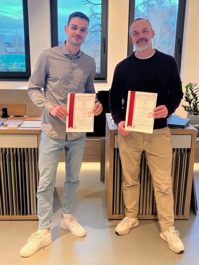 Philipp Hinz und Kai Forg stehen in einem Raum, lächeln in die Kamera und halten Urkunden in den Händen. Beide tragen helle Turnschuhe und Freizeithosen. Hinter ihnen stehen zwei Dokumentenablagen und große Fenster, durch die man einen schwachen Blick nach draußen erhaschen kann. Die Beleuchtung wirkt warm und natürlich. Philipp Hinz und Kai Forg stehen in einem Raum, lächeln in die Kamera und halten Urkunden in den Händen. Beide tragen helle Turnschuhe und Freizeithosen. Hinter ihnen stehen zwei Dokumentenablagen und große Fenster, durch die man einen schwachen Blick nach draußen erhaschen kann. Die Beleuchtung wirkt warm und natürlich..